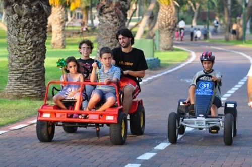 משפחה נוסעת על מכונית פדלים חשמלית אדומה ליד ילד שנוסע על מכונית פדלים חשמלית כחולה